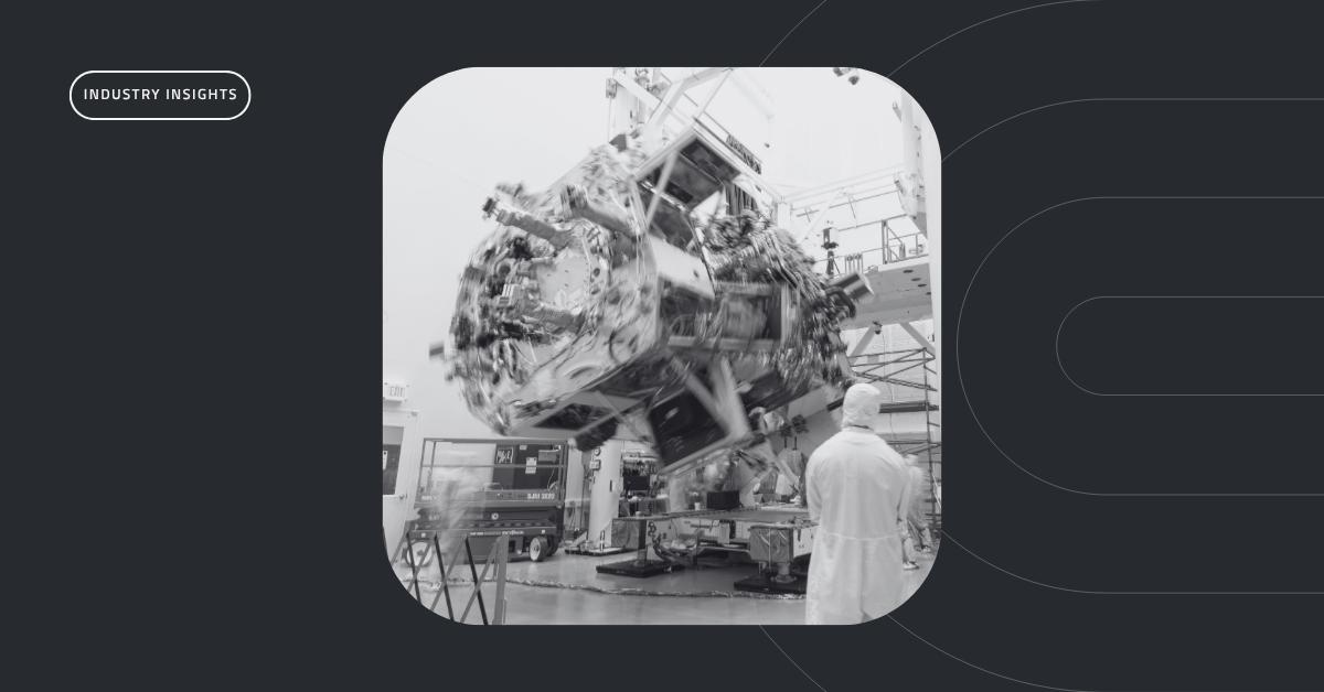 An aerospace technician in a cleanroom suit observing a spacecraft component being lifted in a high-bay assembly facility.